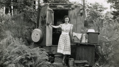 A vintage 1940s black and white photo of woman standing in front of an old caravan wearing a patterned day dress.